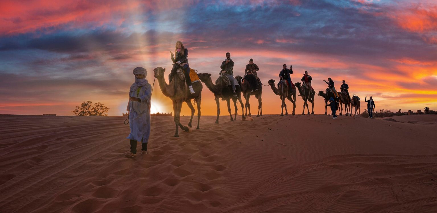 Sahara, Morocco. October 10, 2021. Male guide leads camels caravan with tourists going through the sand in sahara desert during sunset, Tourists are riding camels in desert